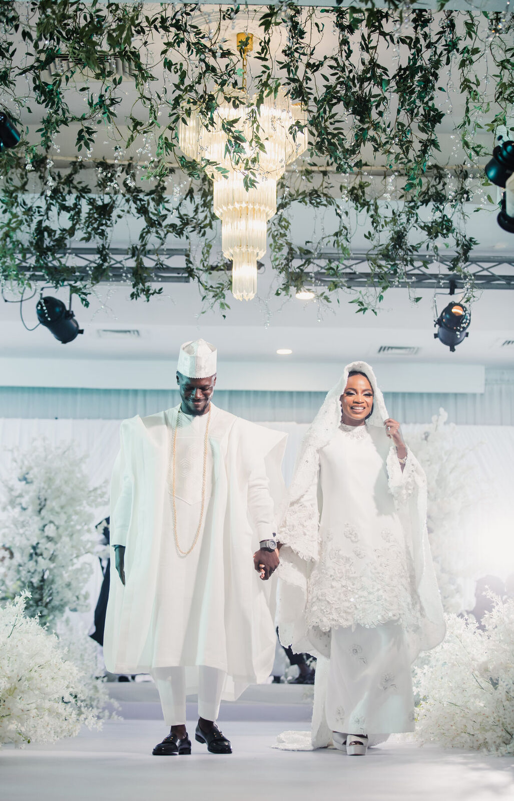 A couple in white agbada and gele walking through a flower-filled hall